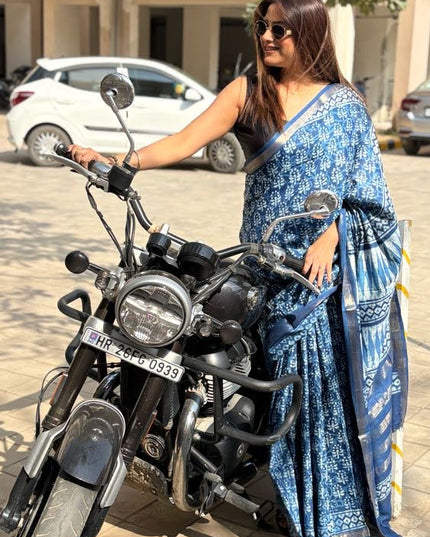 Woman in a blue saree standing next to a motorcycle on a paved area with cars in the background.