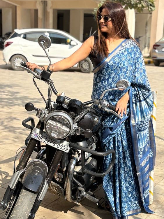 Woman in a blue saree standing next to a motorcycle on a paved area with cars in the background.