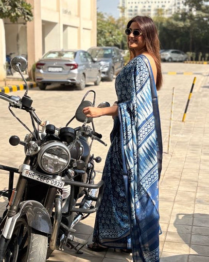 Woman in a blue saree standing next to a motorcycle on a street.